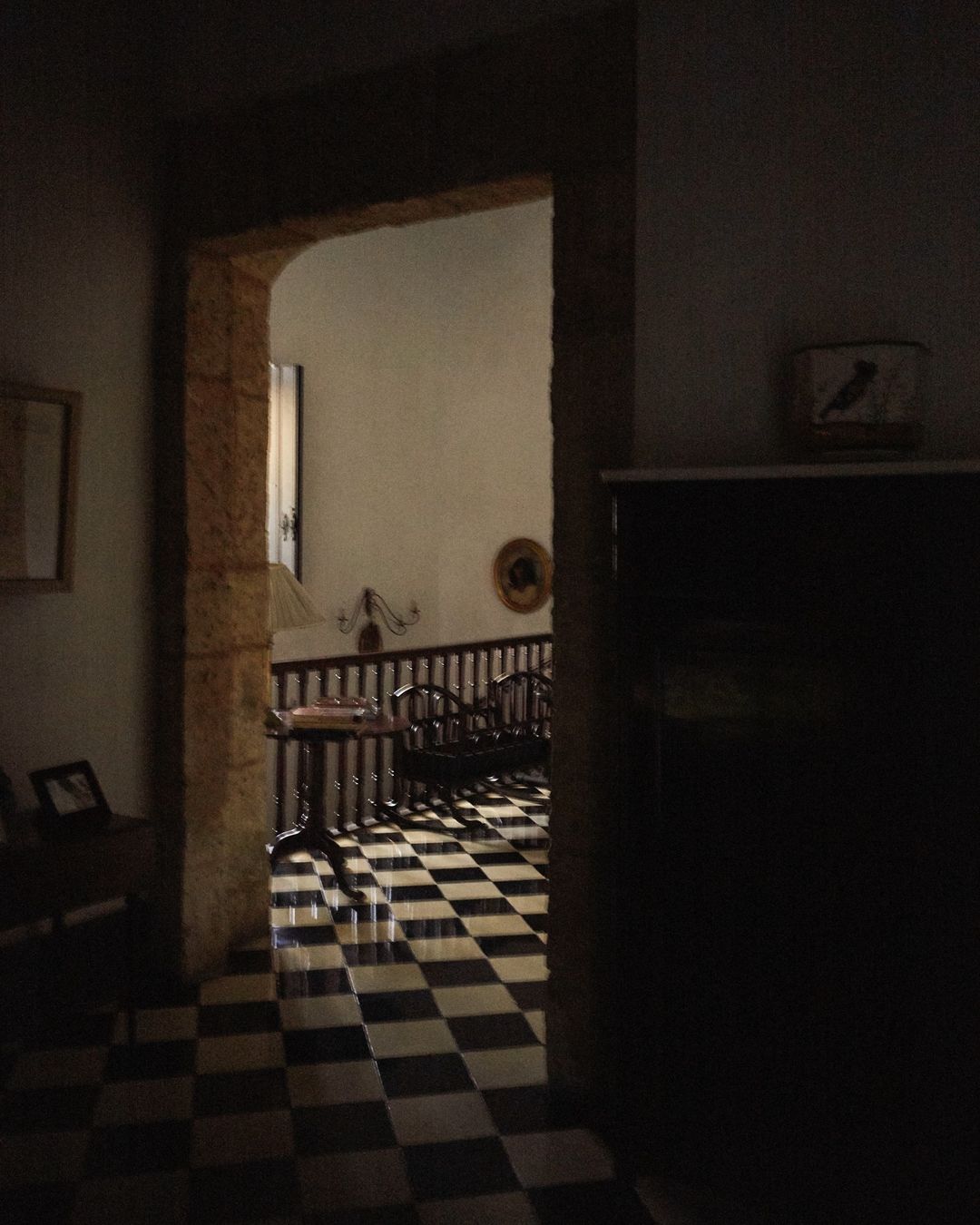 Interior view through arched doorway featuring black-and-white checkered tile flooring, wooden railing, table, and hints of natural light.