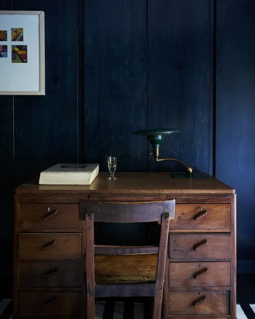 Dark wood desk featuring a lamp, chair, book, and glass set against a dark blue paneled wall with an abstract painting.