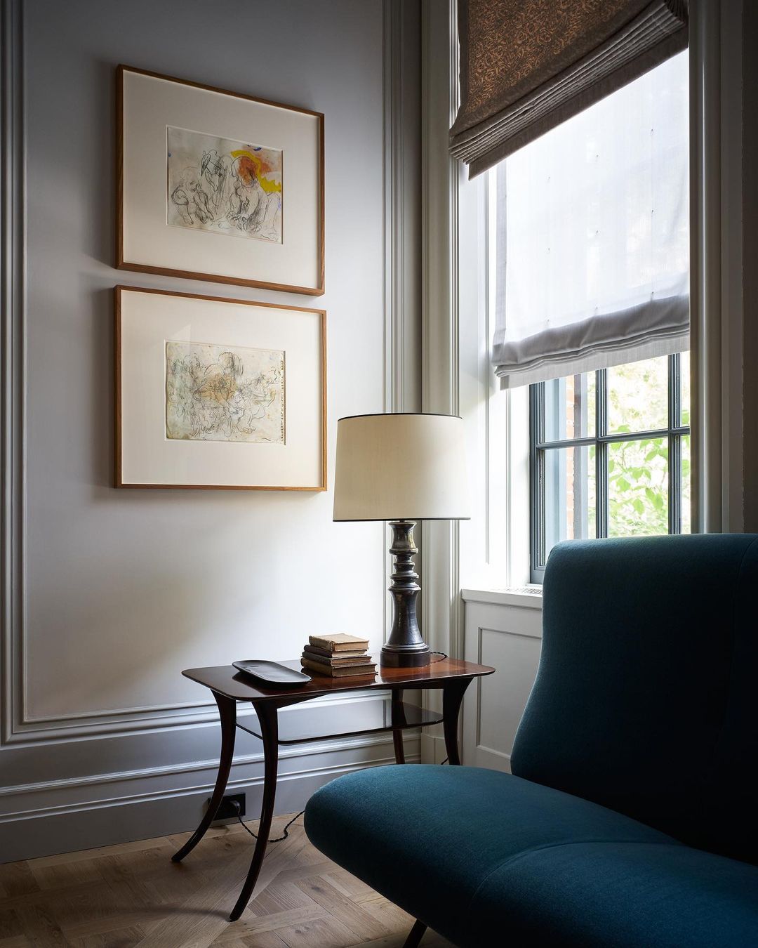 Living room interior featuring a blue chair, a side table with books and a lamp, and artwork hanging on a gray wall by a window.