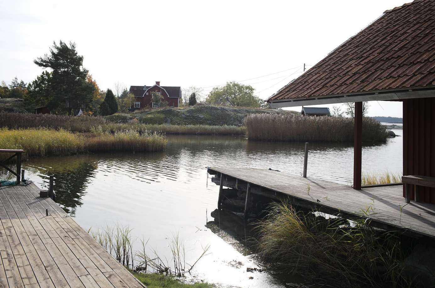 Scenic view of calm water reflecting a red house and lush greenery, with a wooden dock in the foreground on a bright day.