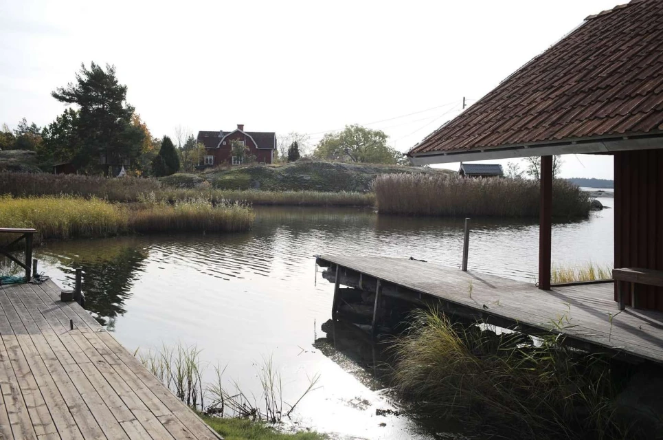 Scenic view of calm water reflecting a red house and lush greenery, with a wooden dock in the foreground on a bright day.