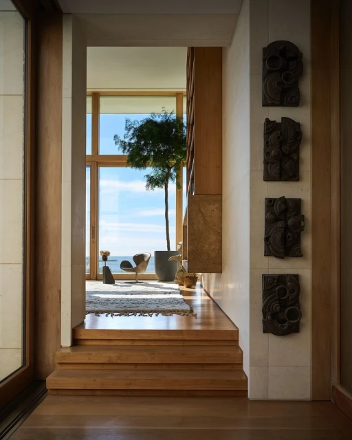 Modern home interior featuring wooden stairs leading to a living area with ocean view, a potted tree, and artistic wall decor.