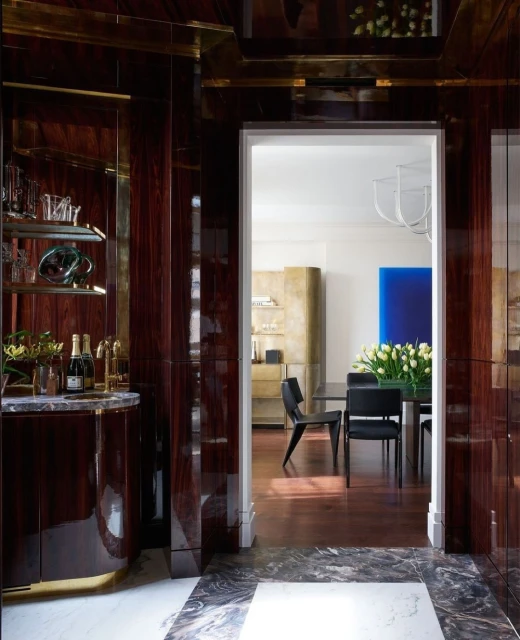 Elegant interior featuring a dark wood bar area with glassware and bottles, leading into a dining space with modern chairs and artwork.