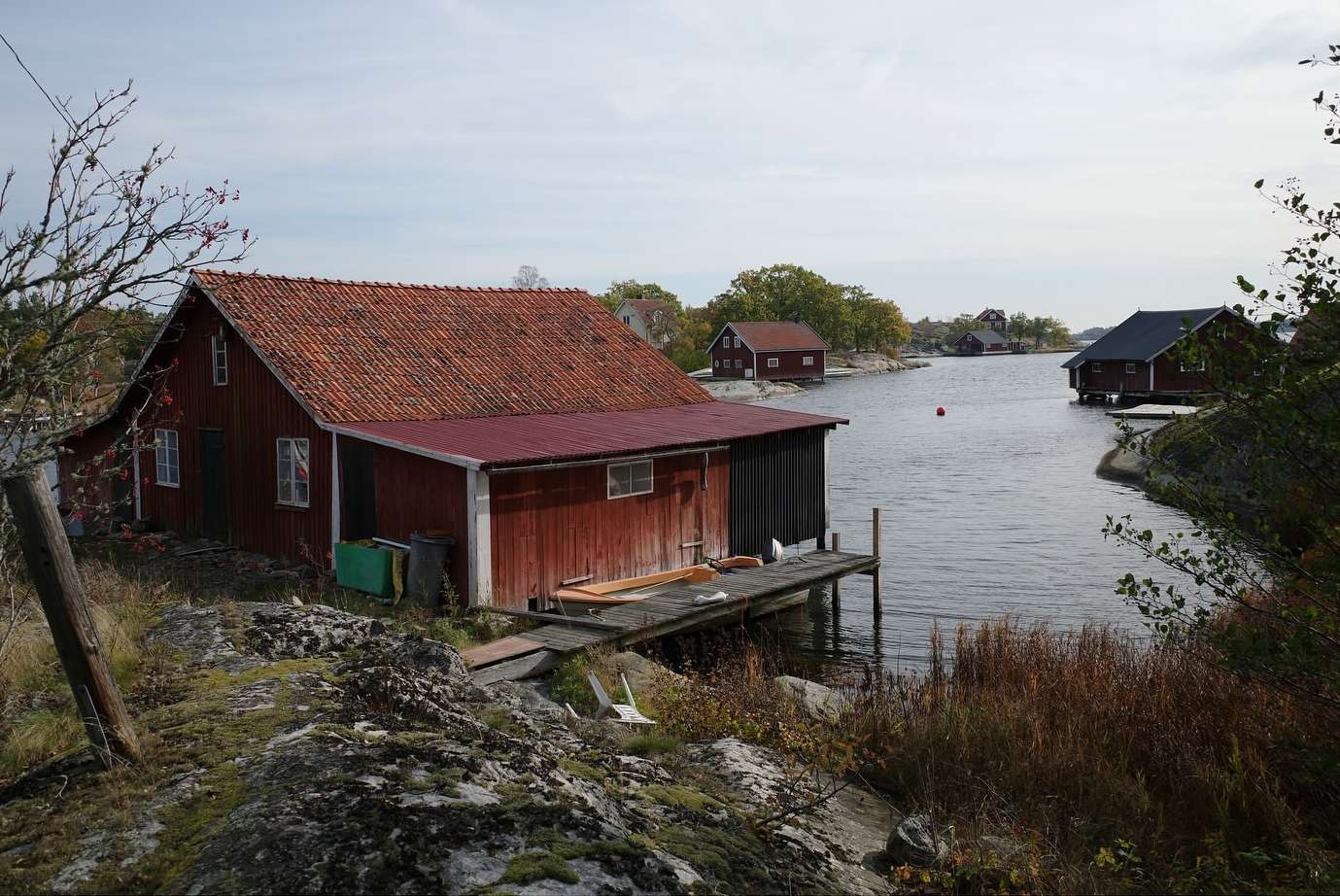 Rural landscape featuring red wooden boathouses along a tranquil riverbank under a cloudy sky, creating a scenic waterscape.