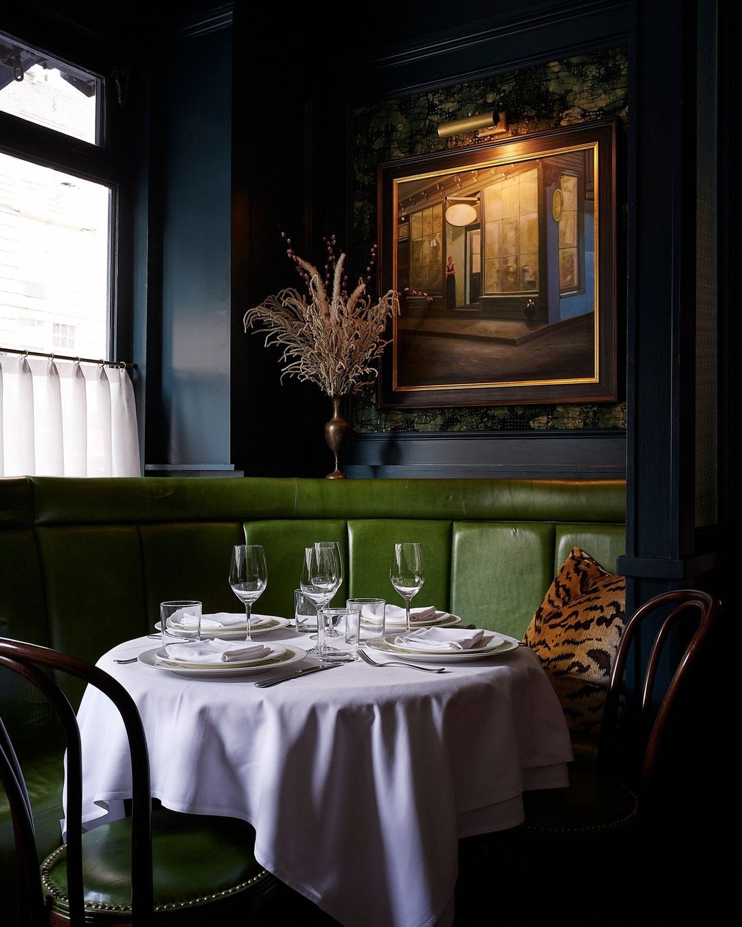 Elegant restaurant interior with a white-clothed table set for four, surrounded by dark green booth seating and dim lighting.