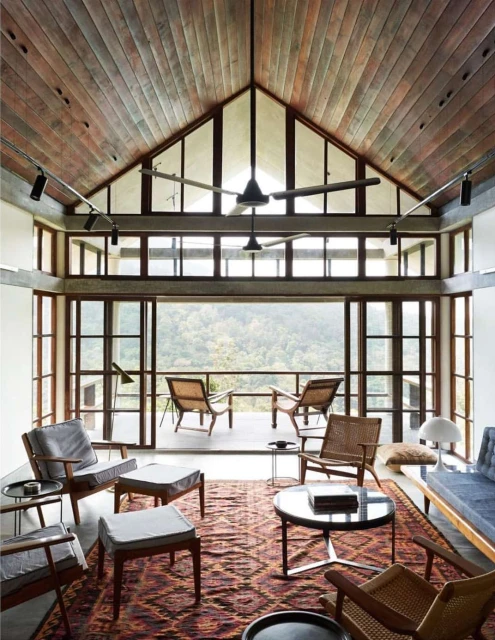 Living room interior featuring mid-century modern furniture, patterned rug, wood ceiling, and large windows with a view of green trees.