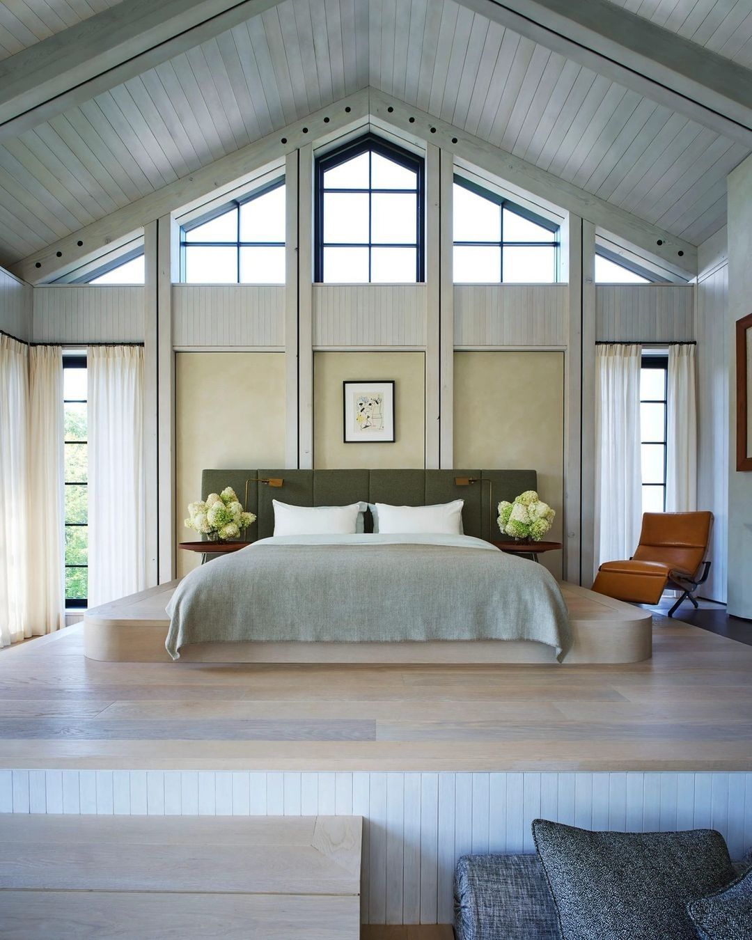 Bedroom interior featuring a bed on a platform, flanked by side tables with white flowers, beneath a high ceiling with skylight windows.