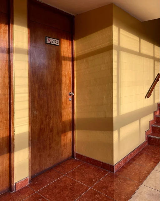 A wooden door with the number 232 on a beige-painted wall in a hallway with red tile floors and stairs with a warm, golden light.