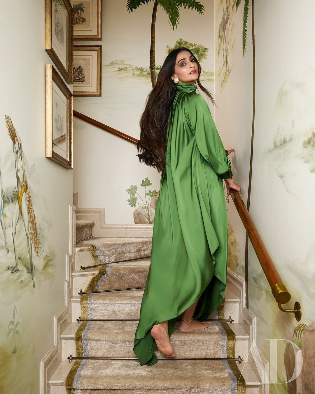 Elegant adult female model in a green satin dress poses on a stairway in an indoor setting with painted walls and wooden handrail.