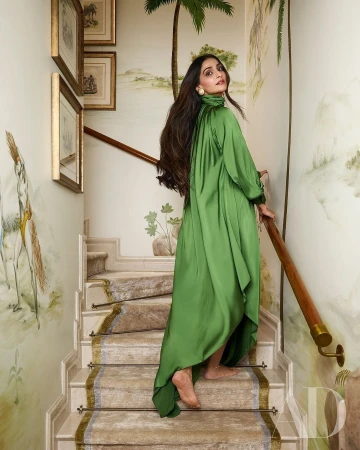 Elegant adult female model in a green satin dress poses on a stairway in an indoor setting with painted walls and wooden handrail.