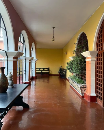 Indoor corridor with brown tile floor, yellow walls, arched doorways, hanging light, and antique table, creating a classic interior.