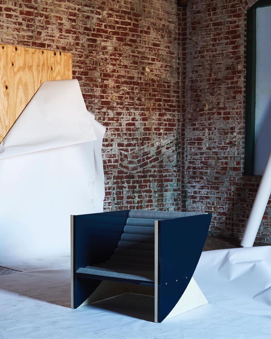 Modern blue accent chair with a gray upholstered seat and backrest, set against a brick wall and draped white fabric in a studio.
