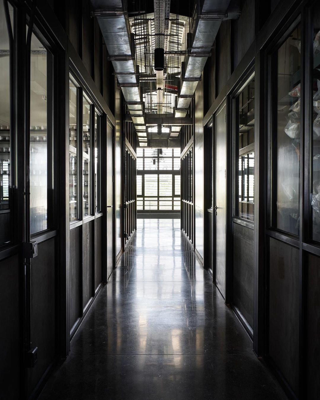 A long indoor corridor with windows, doors, and metal pipes running across the ceiling. The perspective is straight down the hall.