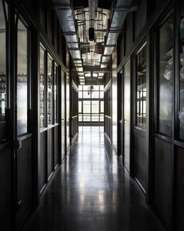 A long indoor corridor with windows, doors, and metal pipes running across the ceiling. The perspective is straight down the hall.