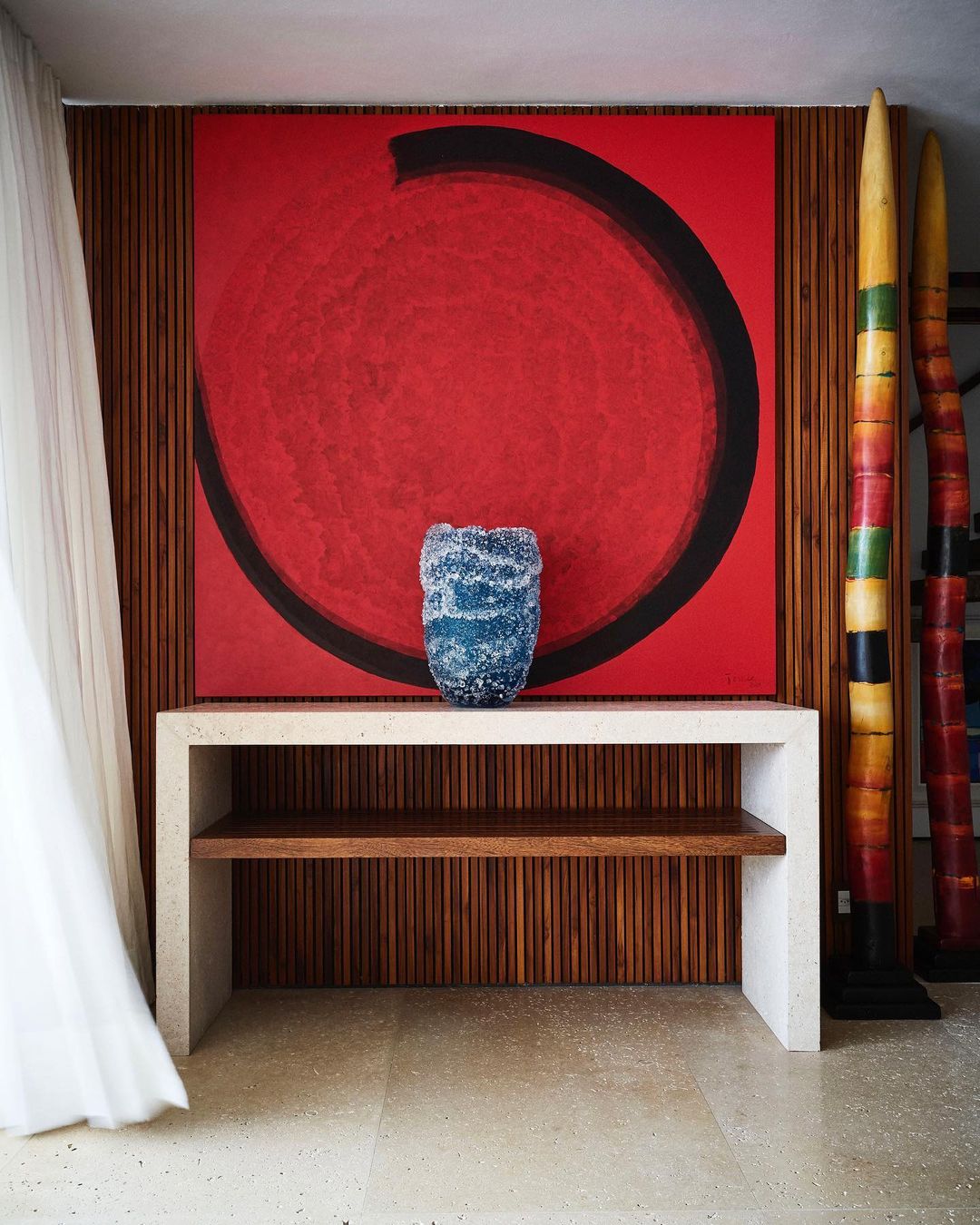 Interior featuring a red abstract painting above a light-colored console table with a blue glass vase in a brightly lit indoor space.