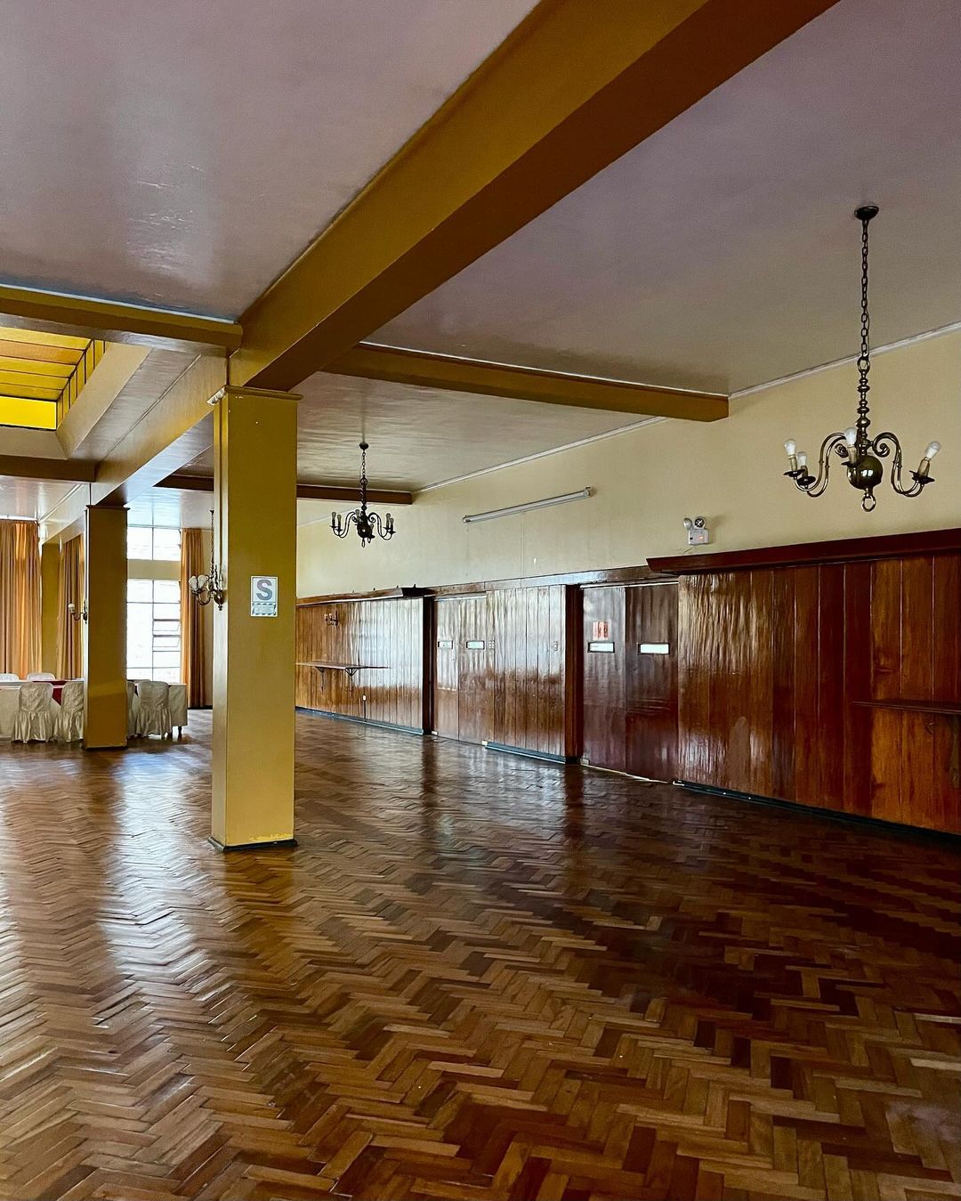 Spacious indoor venue featuring herringbone wood floors, paneled walls, decorative lighting, and high ceilings with supporting columns.