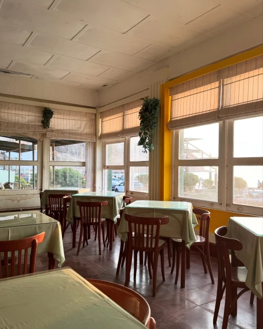 Sunlit restaurant interior featuring empty wooden tables and chairs with pale green tablecloths, and large windows with slatted blinds.