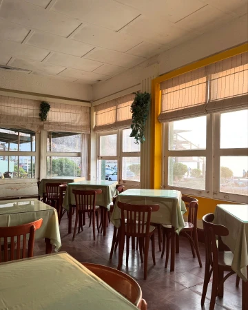 Sunlit restaurant interior featuring empty wooden tables and chairs with pale green tablecloths, and large windows with slatted blinds.