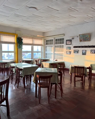 Interior of a restaurant with chairs and tables covered in cloths, illuminated by light coming through the windows.