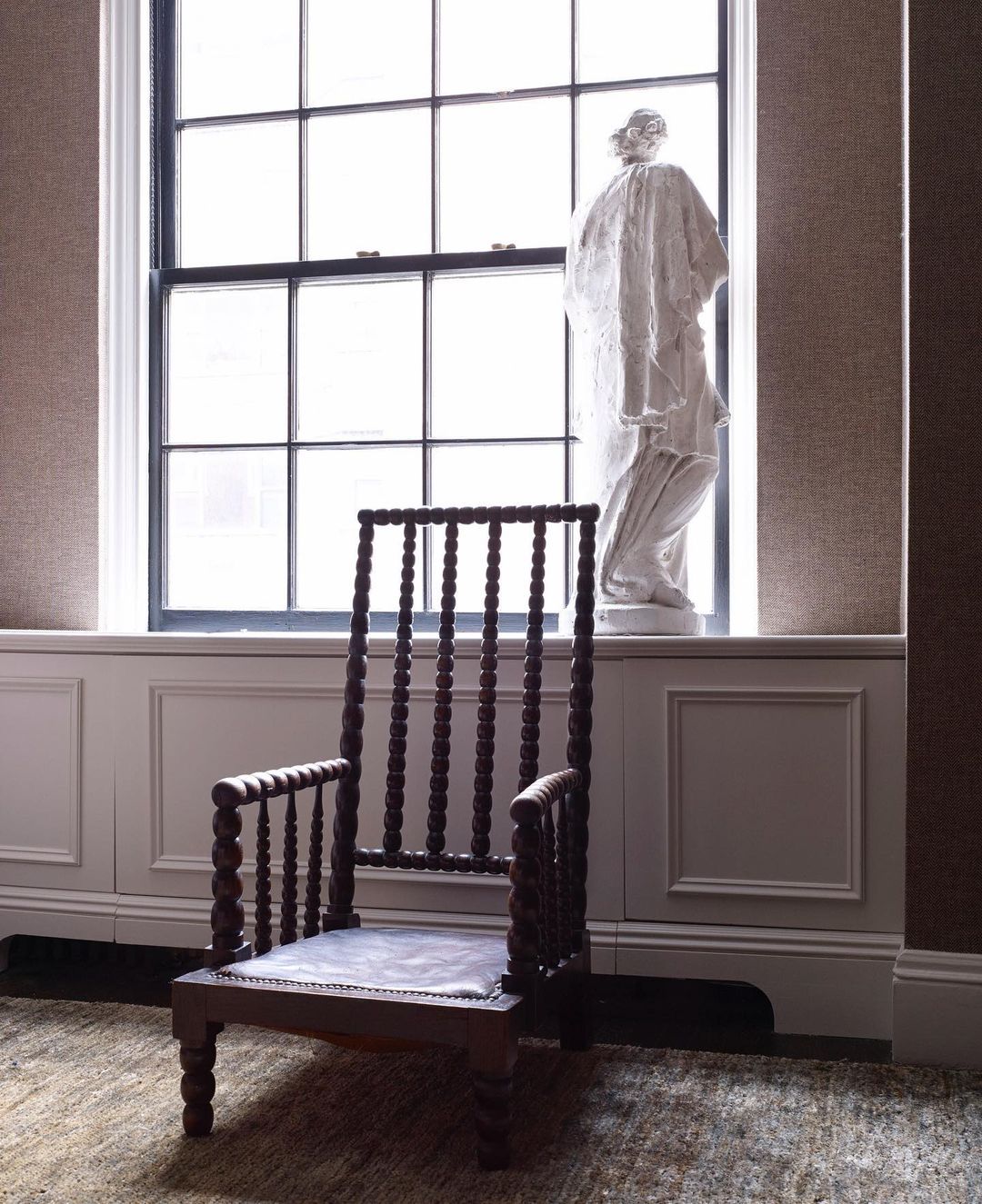 Interior shot of a dark wood bobbin chair near a window with a classical statue on the window sill, against a neutral backdrop.