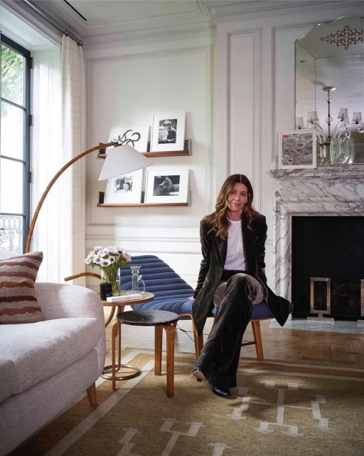 Adult female sits on a blue chair in a bright living room with a fireplace, modern art, and neutral toned furniture. Interior shot.