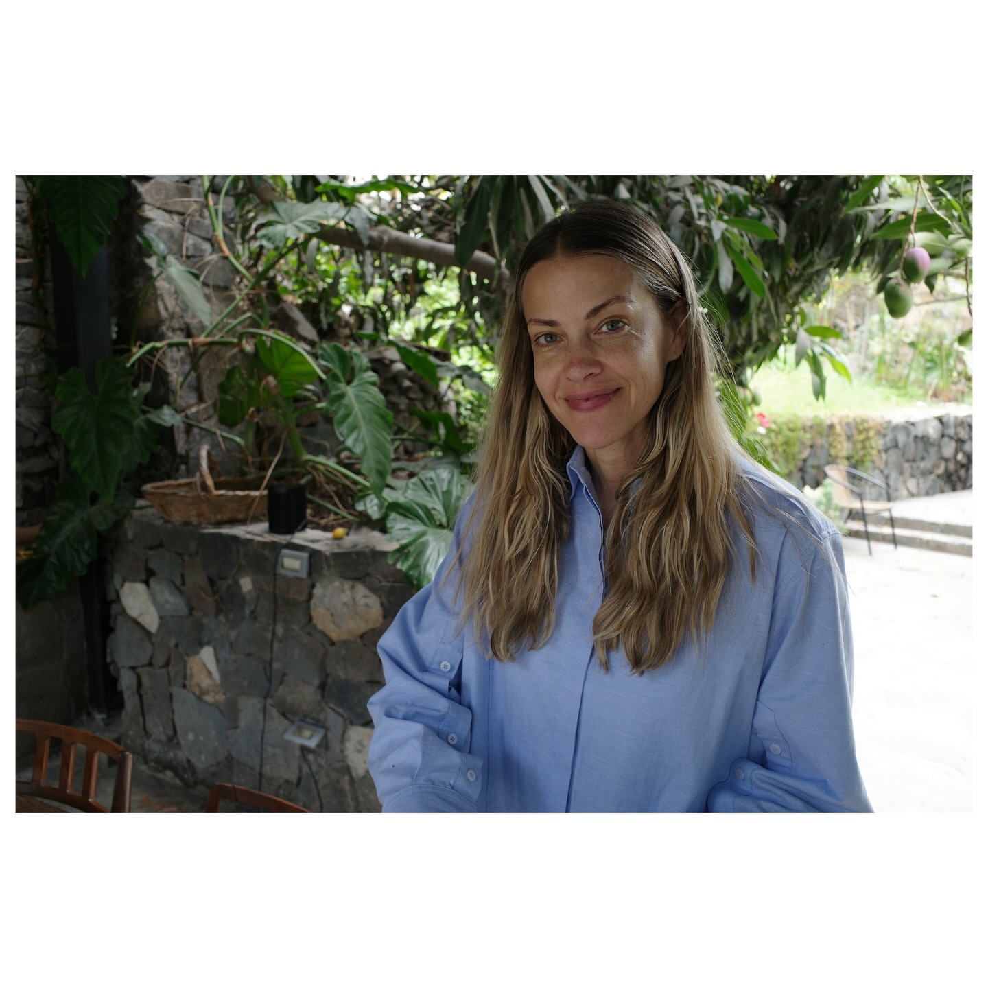 Portrait of a woman smiling, wearing a blue button-down shirt, posing outdoors with lush green foliage and a stone wall in the background.