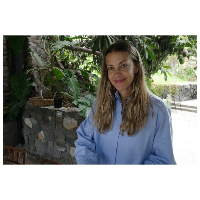 Portrait of a woman smiling, wearing a blue button-down shirt, posing outdoors with lush green foliage and a stone wall in the background.