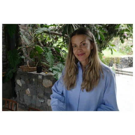 Portrait of a woman smiling, wearing a blue button-down shirt, posing outdoors with lush green foliage and a stone wall in the background.