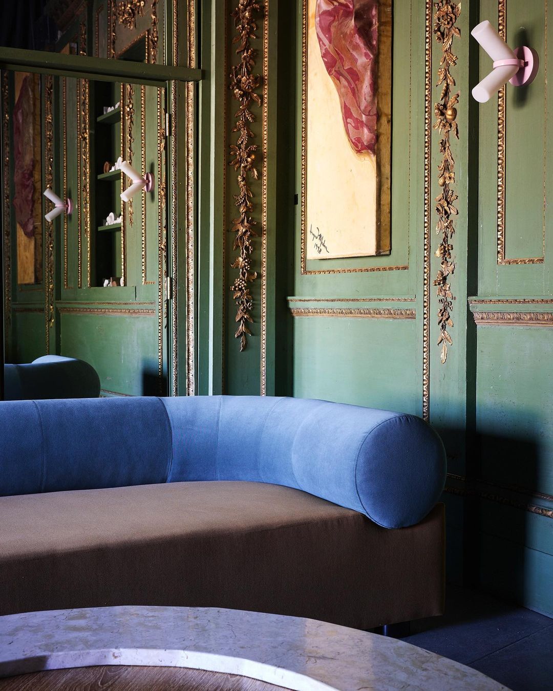 Ornate interior with green walls, gold trim, and a blue-and-brown curved sofa, accented by modern sconces and a painting.