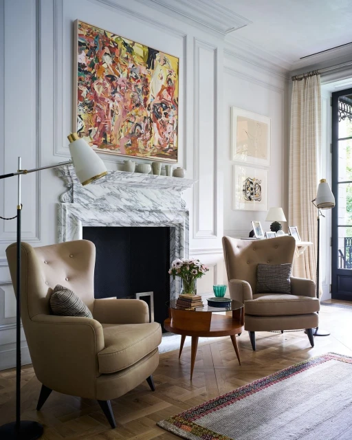 Living room interior featuring two beige armchairs, a round coffee table with flowers, a fireplace with art, and light hardwood floors.