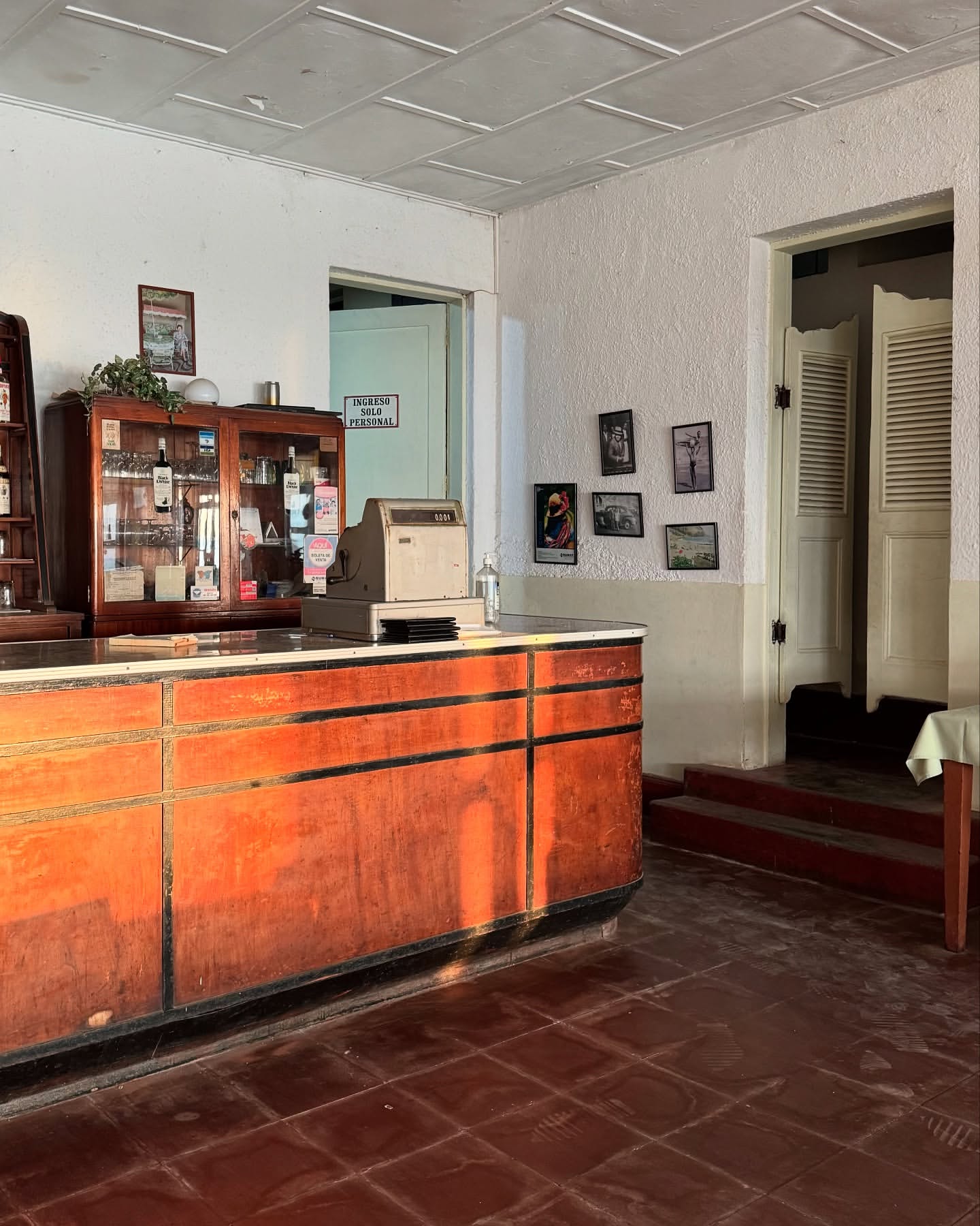 Interior shot of a vintage room featuring a wood counter, display case, cash register, tiled floor, and textured white walls.