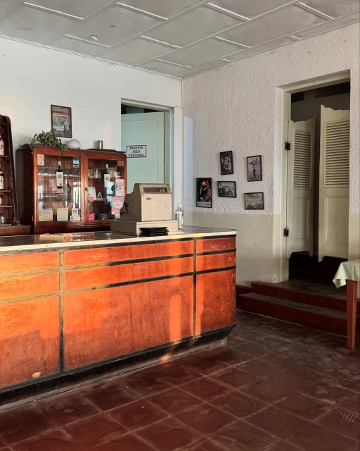 Interior shot of a vintage room featuring a wood counter, display case, cash register, tiled floor, and textured white walls.