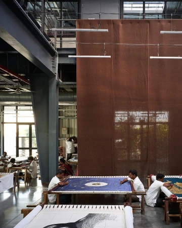People in a factory work on embroidery projects, seated at long tables in a large industrial setting with neutral tones and natural lighting.
