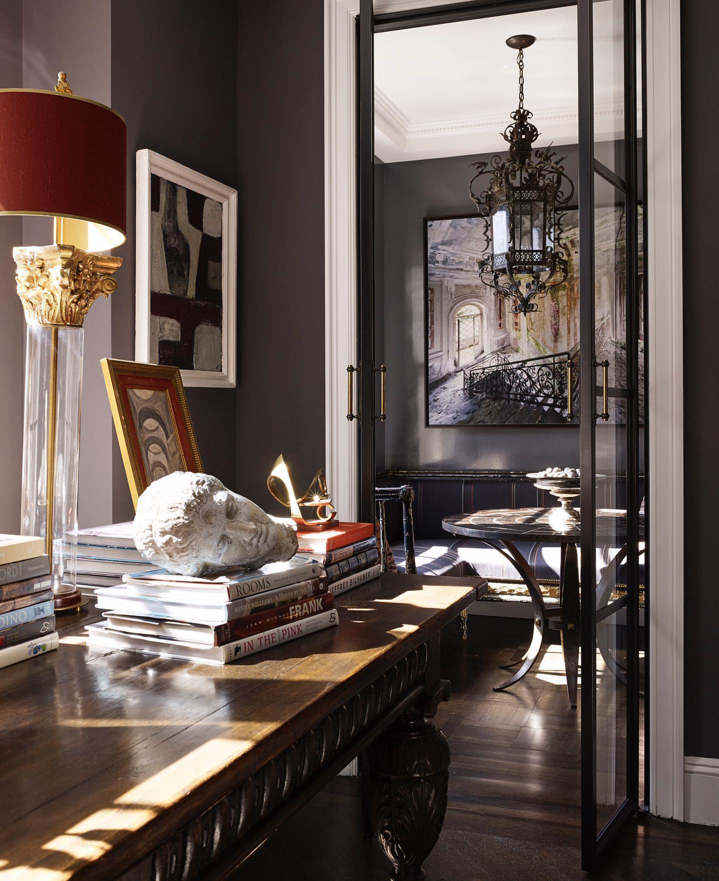 Ornate interior room with antique-style desk topped with books and a bust, illuminated by a classic lamp with a red lampshade.