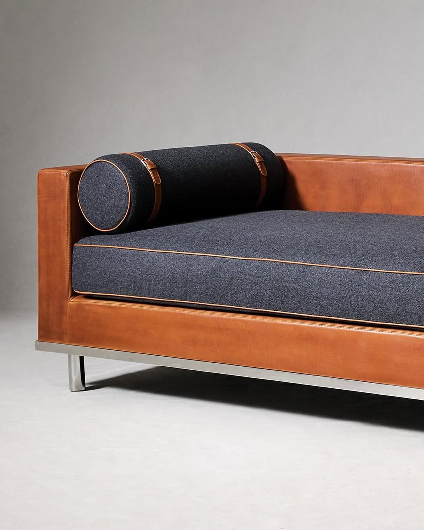 Minimalist tan leather daybed with a bolster pillow featuring dark grey upholstery and tan leather straps, set against a neutral background.