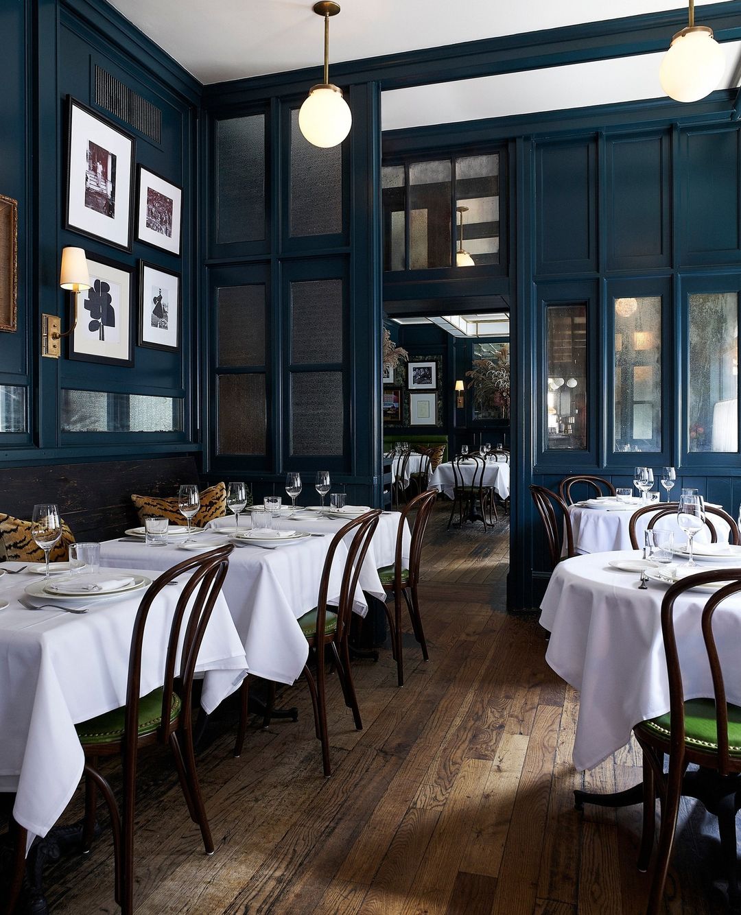 Upscale restaurant interior featuring set tables with white linens, bentwood chairs, hardwood floors, and dark blue wall paneling.