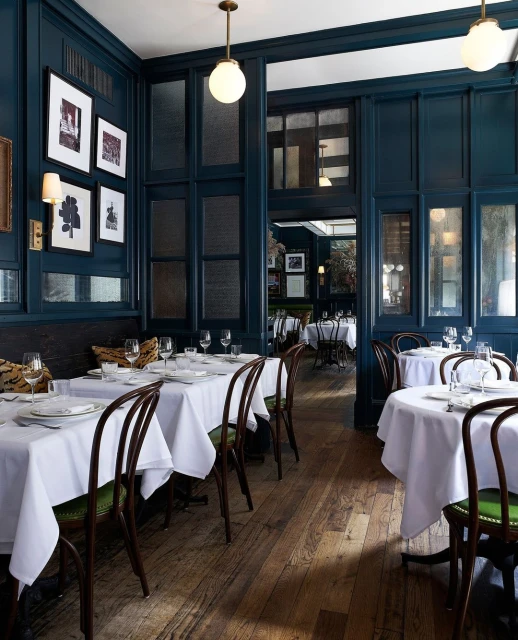 Upscale restaurant interior featuring set tables with white linens, bentwood chairs, hardwood floors, and dark blue wall paneling.