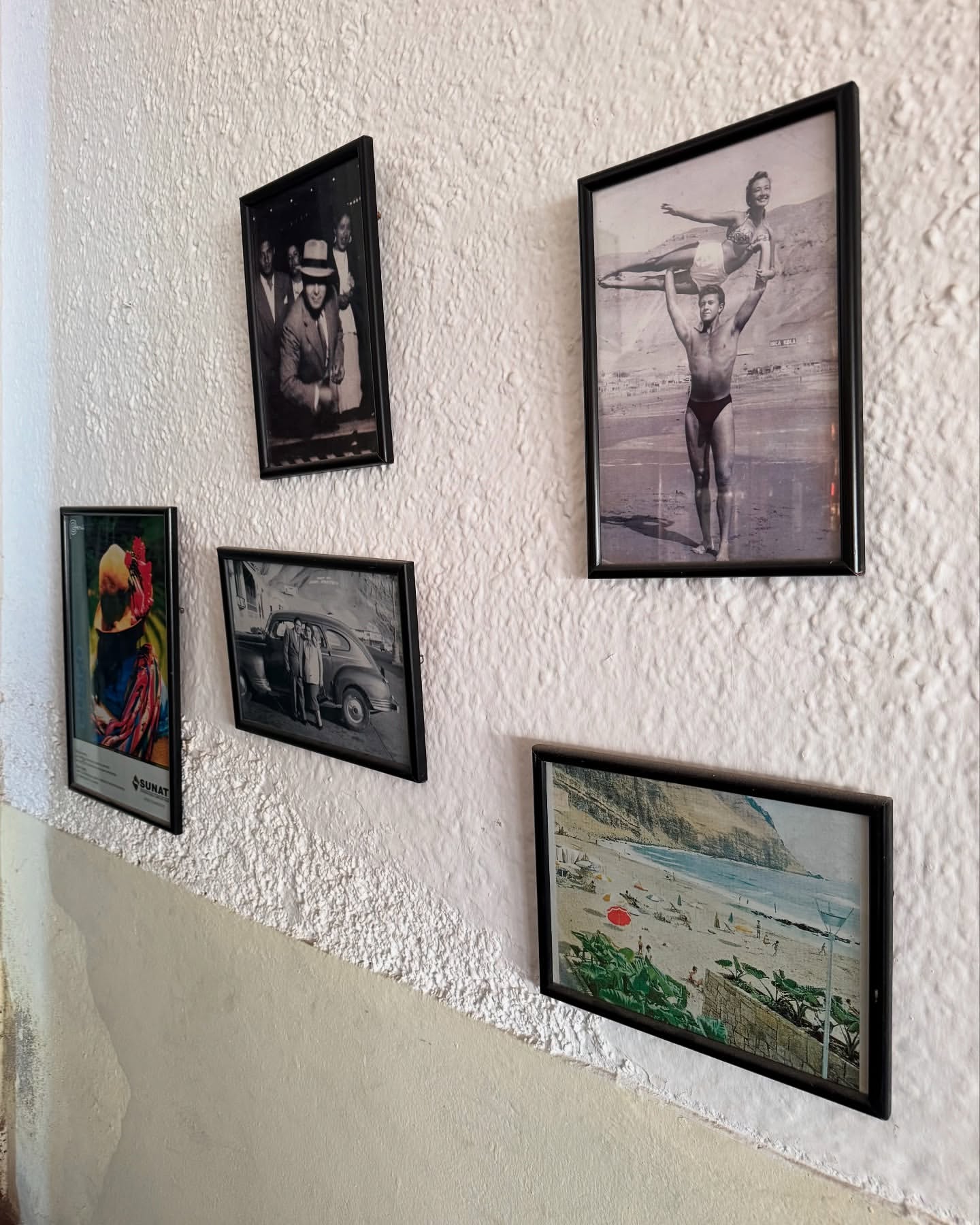 A textured white wall displays five framed photographs and art, including people, vintage cars, and a coastal beach scene in a room.