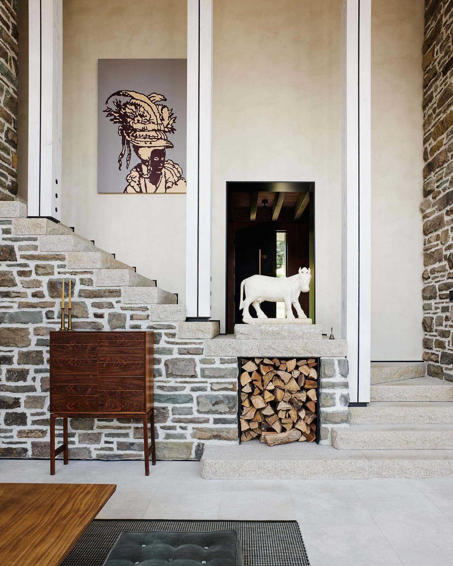 Interior shot of a foyer with stone walls and stairs, featuring a painting, wooden cabinet, sculpture, and stacked firewood.