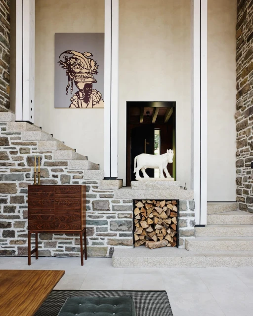 Interior shot of a foyer with stone walls and stairs, featuring a painting, wooden cabinet, sculpture, and stacked firewood.