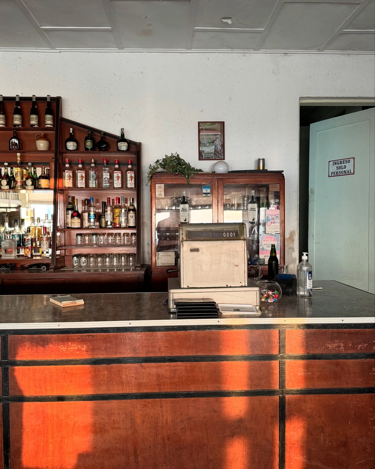 Interior view of a vintage bar with shelves of bottles, a cash register, and a wooden counter in warm light, creating an inviting atmosphere.
