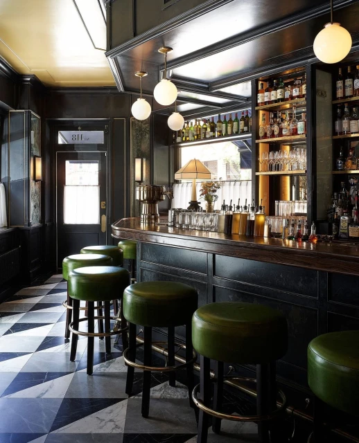 Interior view of a bar with green leather stools, dark wood counter, liquor bottles, and marble tile flooring in a dimly lit setting.