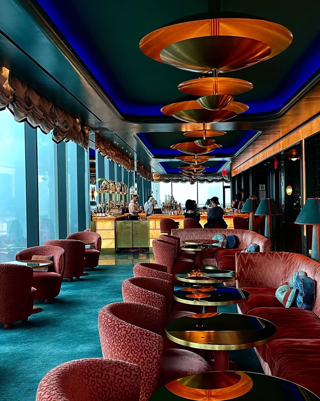 Interior view of a stylish lounge with plush red seating, gold-accented tables, and a well-stocked bar under layered lighting fixtures.