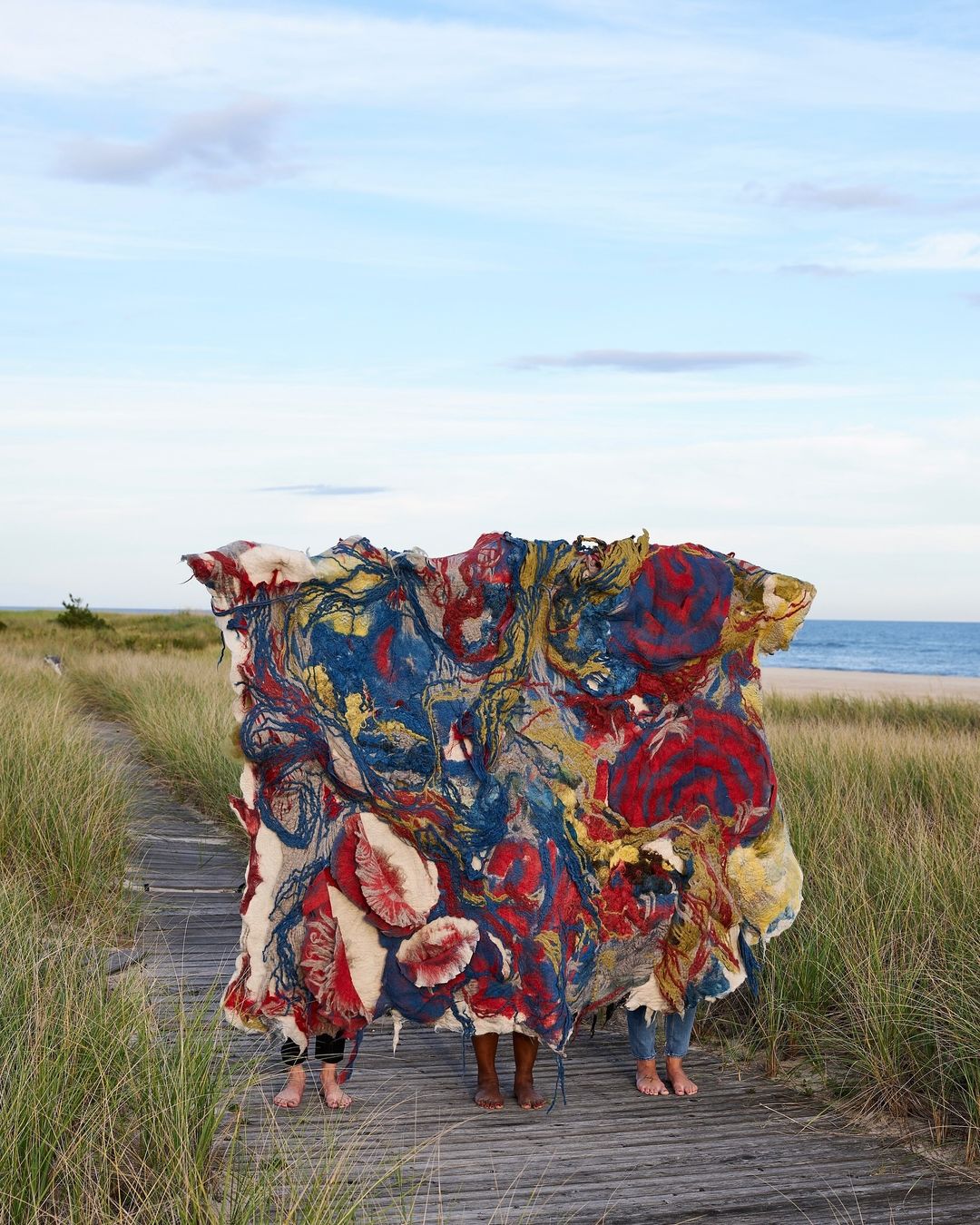 Three people stand on a wooden boardwalk on a beach, concealed by a large, colourful, abstract textile art piece, blue sky above.