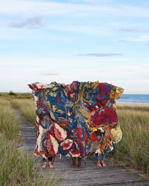 Three people stand on a wooden boardwalk on a beach, concealed by a large, colourful, abstract textile art piece, blue sky above.