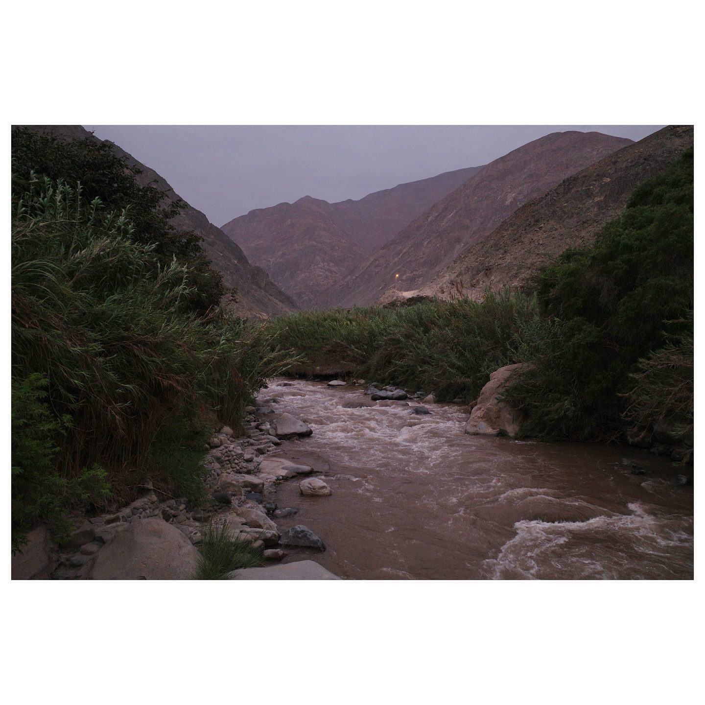 River flows through a valley between mountains with lush green vegetation on the riverbanks on an overcast day with dramatic skies.