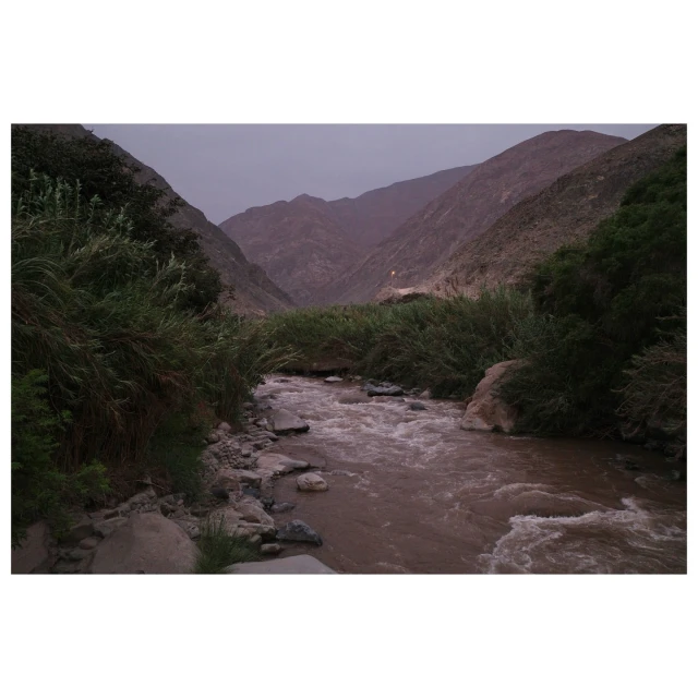 River flows through a valley between mountains with lush green vegetation on the riverbanks on an overcast day with dramatic skies.