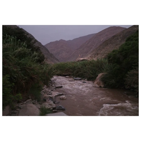 River flows through a valley between mountains with lush green vegetation on the riverbanks on an overcast day with dramatic skies.