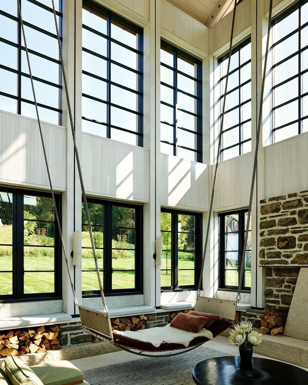 Interior of a sunroom-style living space with a hanging swing chair, stone fireplace, and expansive windows overlooking a green lawn.