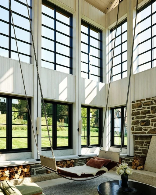 Interior of a sunroom-style living space with a hanging swing chair, stone fireplace, and expansive windows overlooking a green lawn.